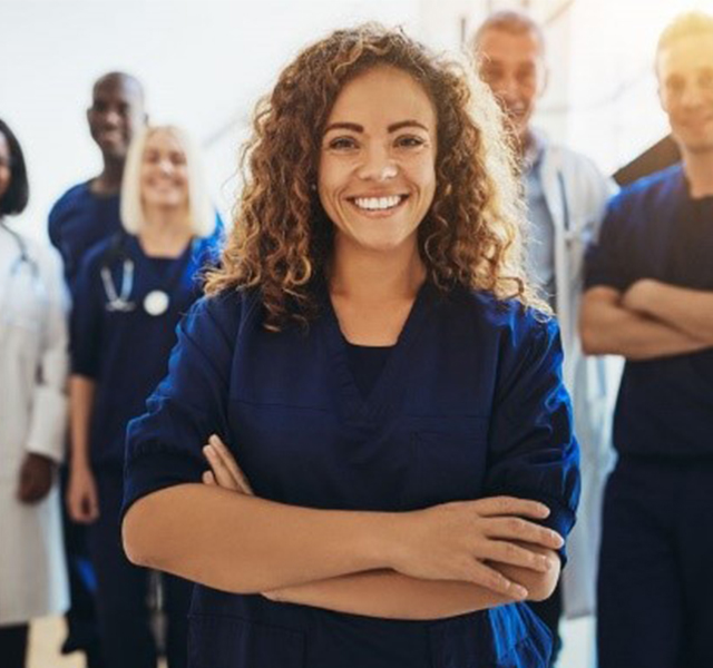 group of six employees wearing blue scrubs and white lab coats. They are standing with their arms folded and smiling.